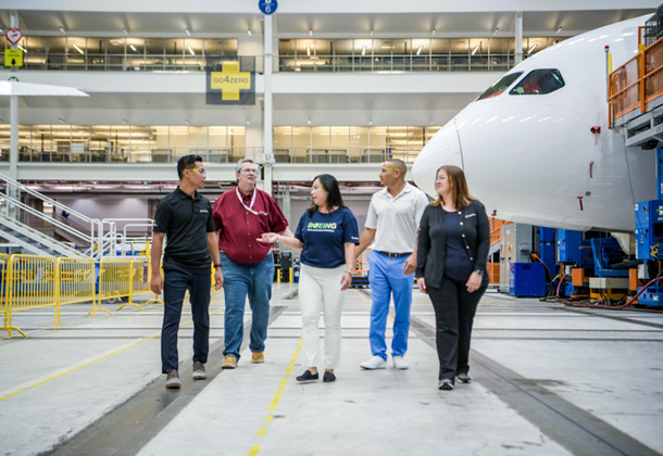 Five Boeing employees of varying ages, genders and races walk across a factory floor, talking with each other. Behind them is the nose of a 787.